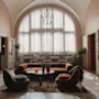 Mukava lounge-tila sisältää pyöreän sohvapöydän, jota ympäröivät modernit tuolit ja sohva. Aurinko paistaa suurista kaari-ikkunoista korostaen kasveja elegantissa huoneessa.