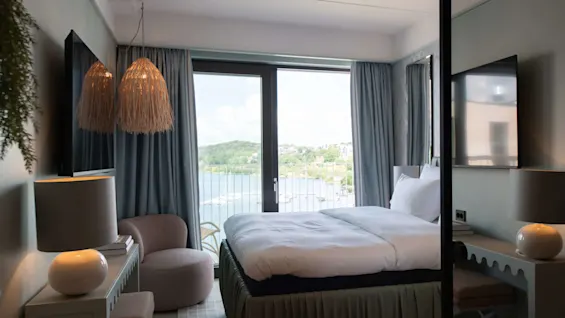 A neatly made bed faces a large window with a view of a serene lake and distant hills. Soft lighting and modern decor accent the cozy bedroom.