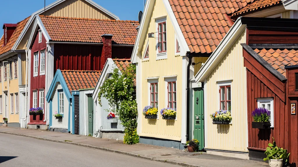 Färgglada trähus med röda, gula och blå fasader kantar en lugn gata. Varje hus har blomlådor fyllda med blommor under fönstren, vilket bidrar till ett pittoreskt område.