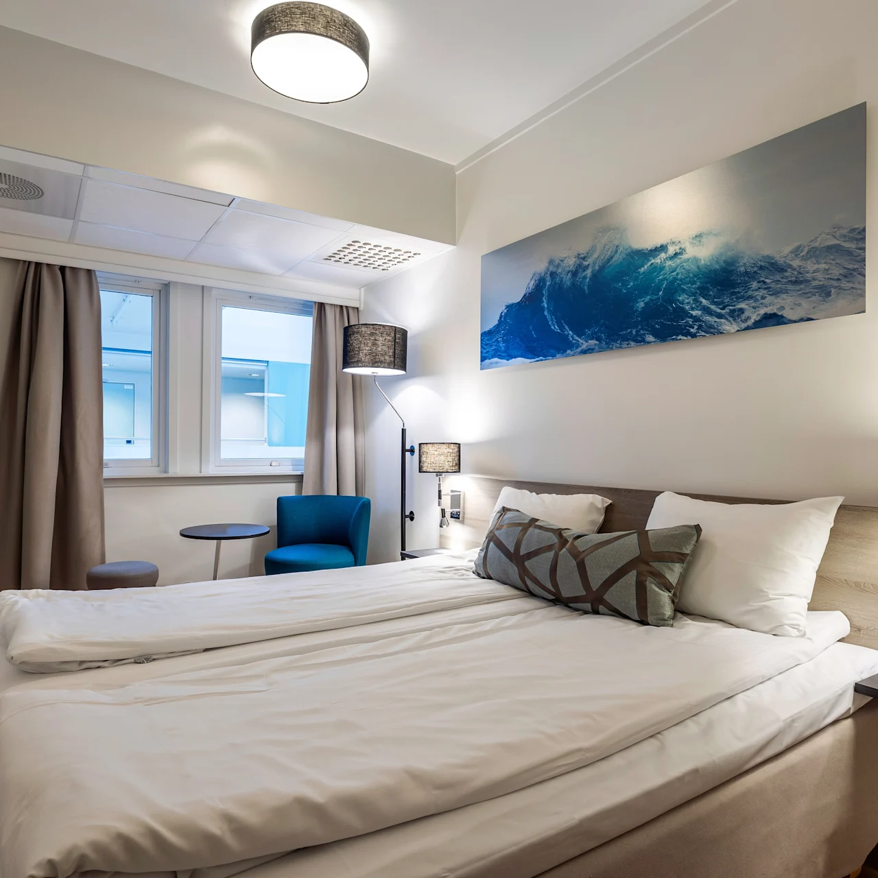 A bed with white linens is positioned in a modern room, flanked by nightstands. A large wave artwork decorates the wall, and a blue chair with a table sits by the window.