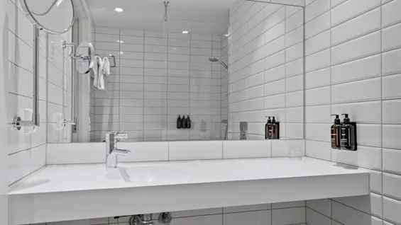 Ett rent, modernt badrum med vita kaklade väggar, en stor spegel över ett handfat och toalettartiklar. En dusch reflekteras i spegeln.