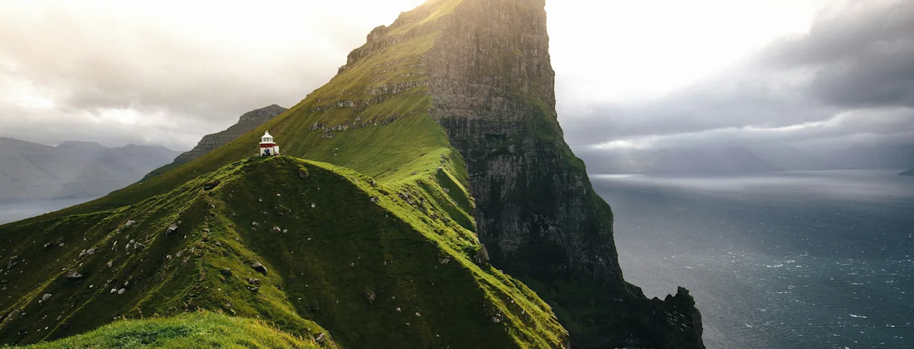 En fantastisk utsikt over en grønn klippe med et lite fyrtårn som skuer ut over havet under en lys himmel på Færøyene.