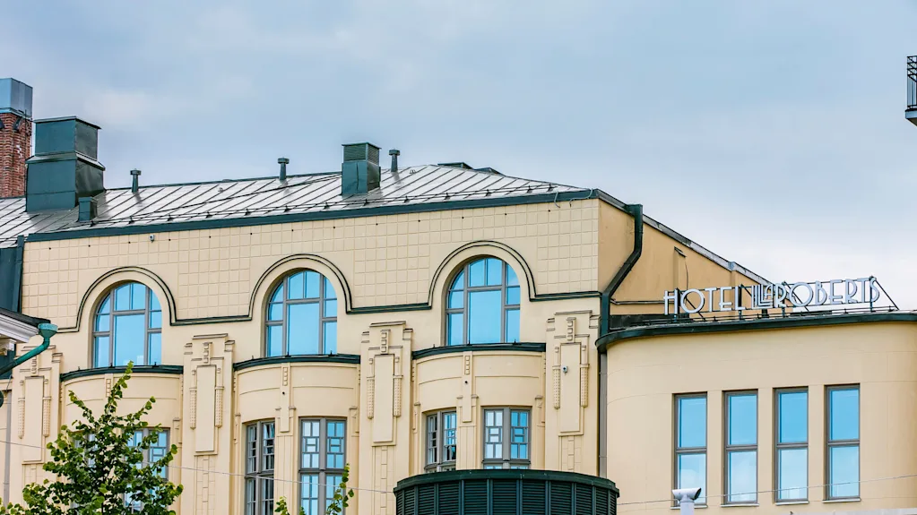 En beige bygning med buede vinduer og et metalltak, med teksten "HOTEL L ROBERTS" på hjørnet, under en overskyet himmel.
