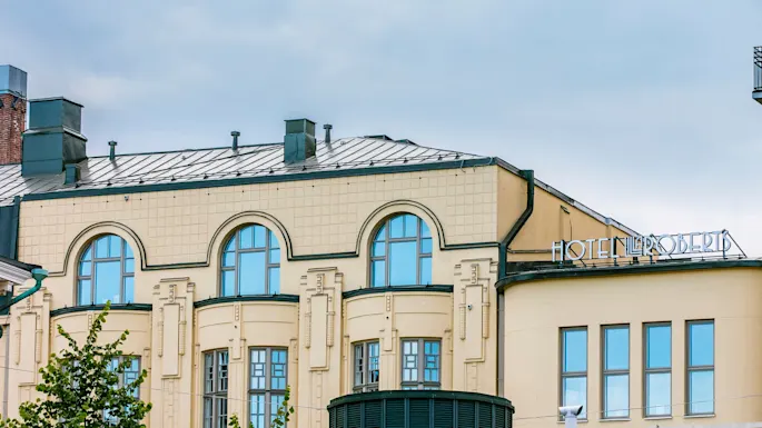 En beige bygning med buede vinduer og et metalltak, med teksten "HOTEL L ROBERTS" på hjørnet, under en overskyet himmel.