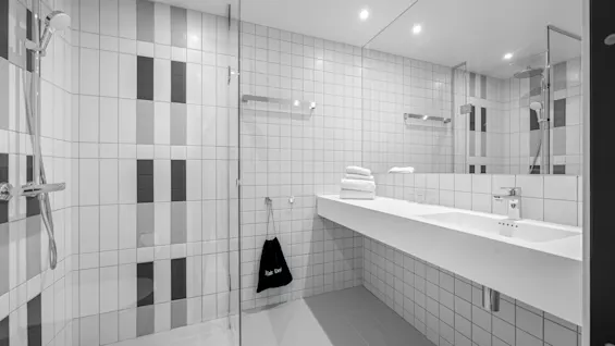 Modern bathroom with a walk-in shower, a long vanity with a sink, and a mirror. A bag labeled "Hair Dry" hangs nearby.