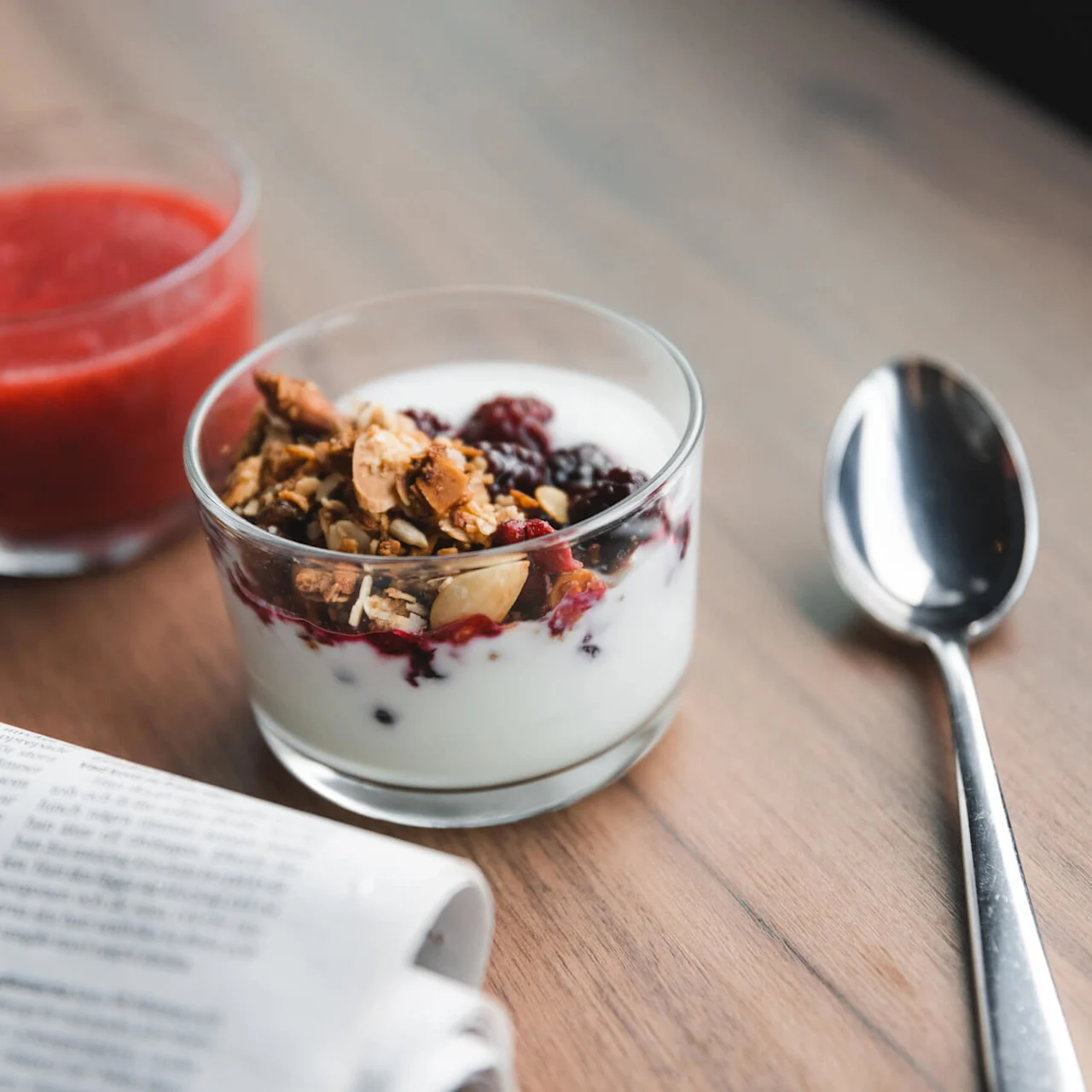 Granola och yoghurt i ett glas bredvid en sked och ett glas med röd juice på ett träbord. En tidning med text på svenska är delvis synlig.