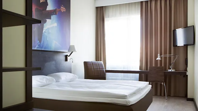 A neatly made bed with white linens stands in a modern hotel room featuring a wall-mounted guitar-themed picture, a small desk with a lamp, and a television.