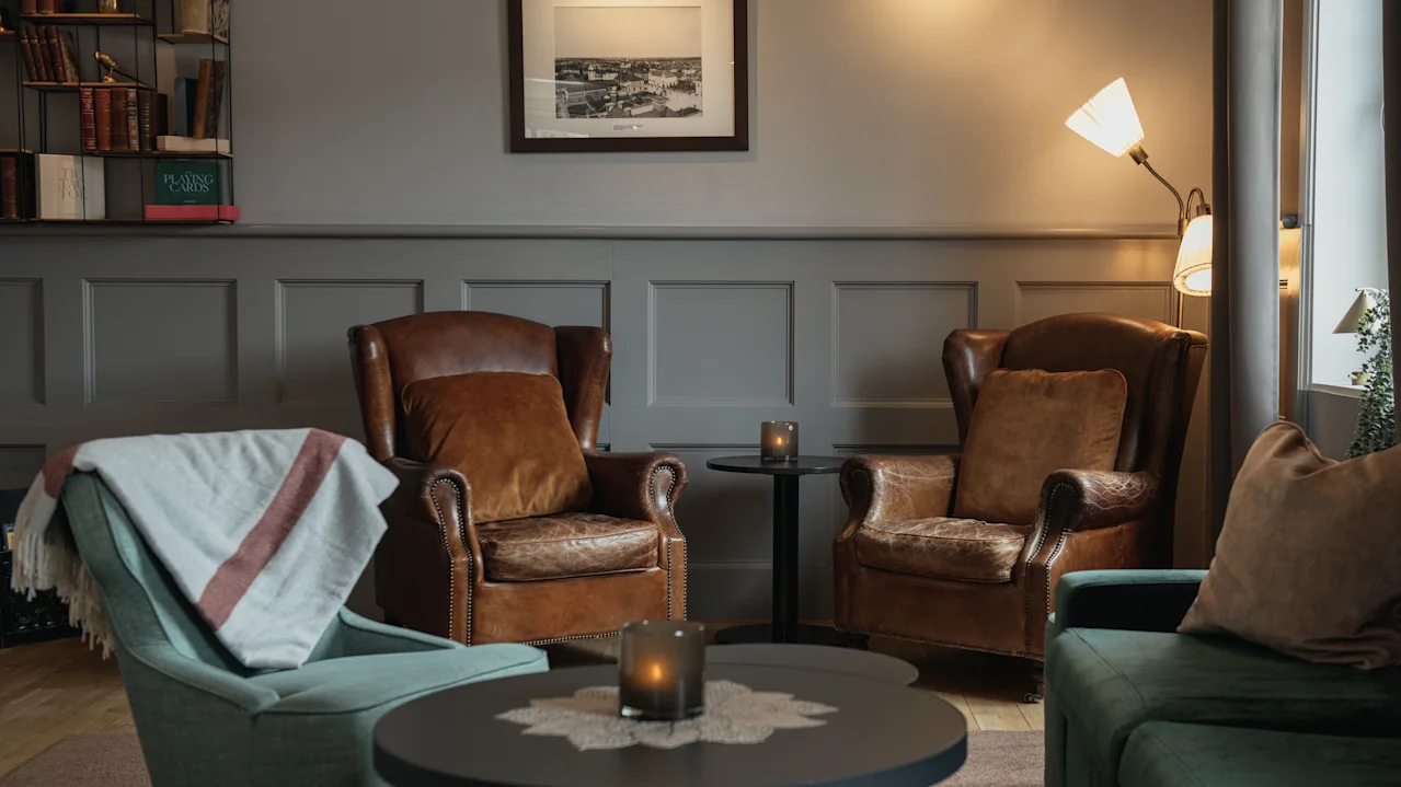 Two leather armchairs sit facing a small round table with candles in a cozy living room. Nearby, a lamp and bookshelves add warmth to the paneled walls.