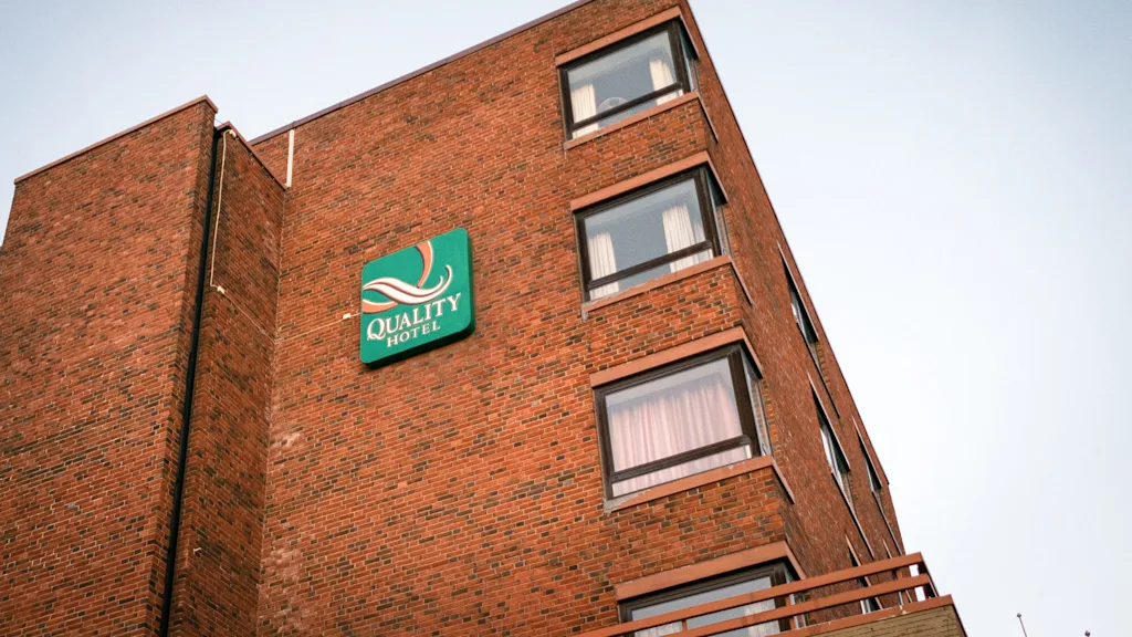 En murstensbygning viser skiltet "QUALITY HOTEL" med store hvide bogstaver på en grøn baggrund, med flere vinduer synlige mod en klar himmel.