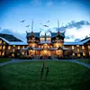 A grand, illuminated wooden mansion with steep roofs stands against a twilight sky. Bats fly overhead, while skeletal arms emerge from the grassy lawn.