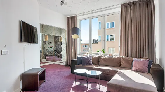 A modern living room with a large corner sofa, flat-screen TV, and an ornate mirror, bathed in natural light from large windows.