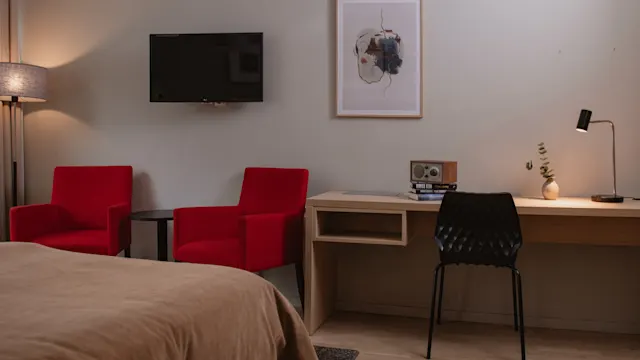 En väggmonterad TV vetter mot två röda fåtöljer bredvid ett skrivbord med en svart stol, lampa, radio och växt i ett modernt hotellrum.
