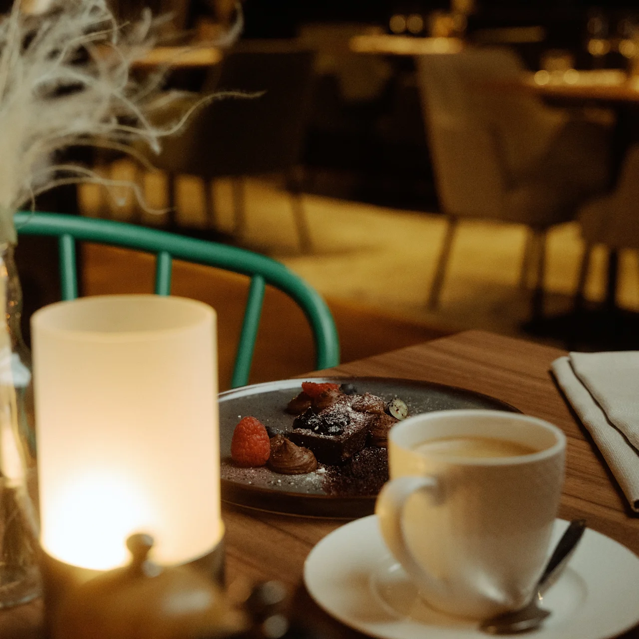 Ett tänt ljus sprider ett varmt sken över ett bord med kaffe, dessert och en vikt servett; omgivet av mjukt belysta, bekväma stolar i en mysig matsalsmiljö.