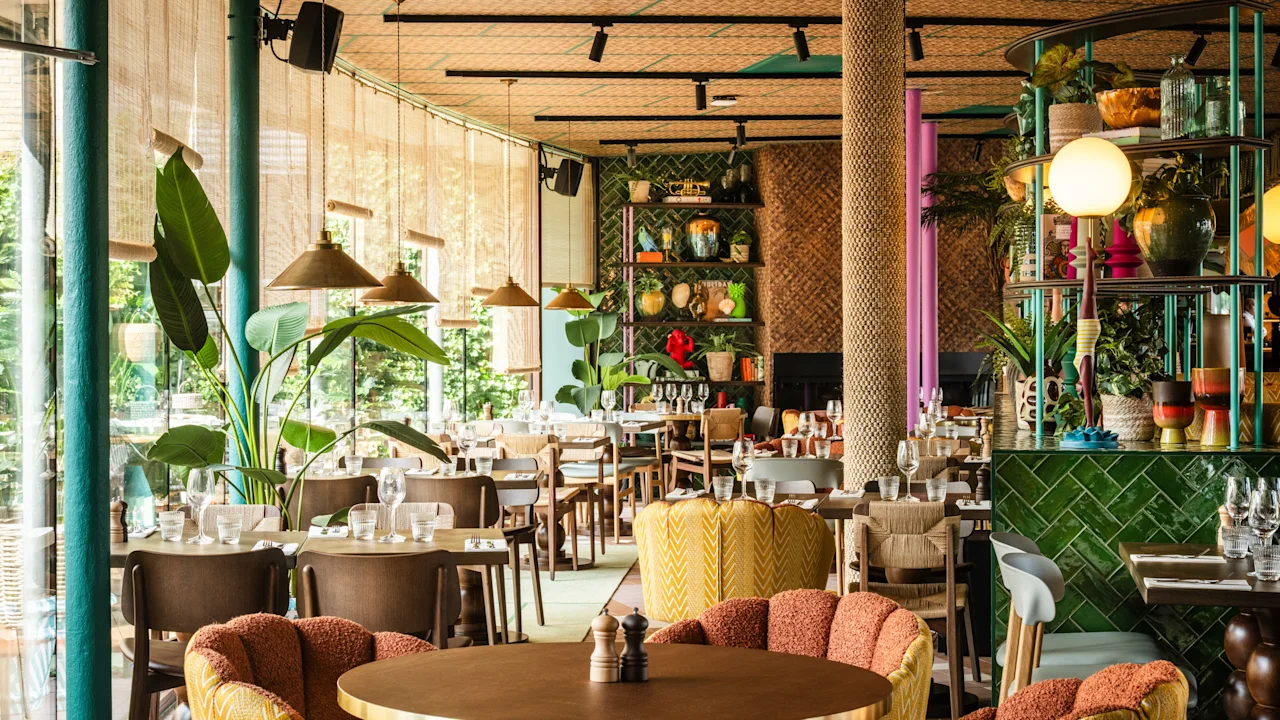 En restaurant med farverige møbler og tropiske planter indenfor, borde dækket med glas og bestik, omgivet af store vinduer der lukker naturligt lys ind.