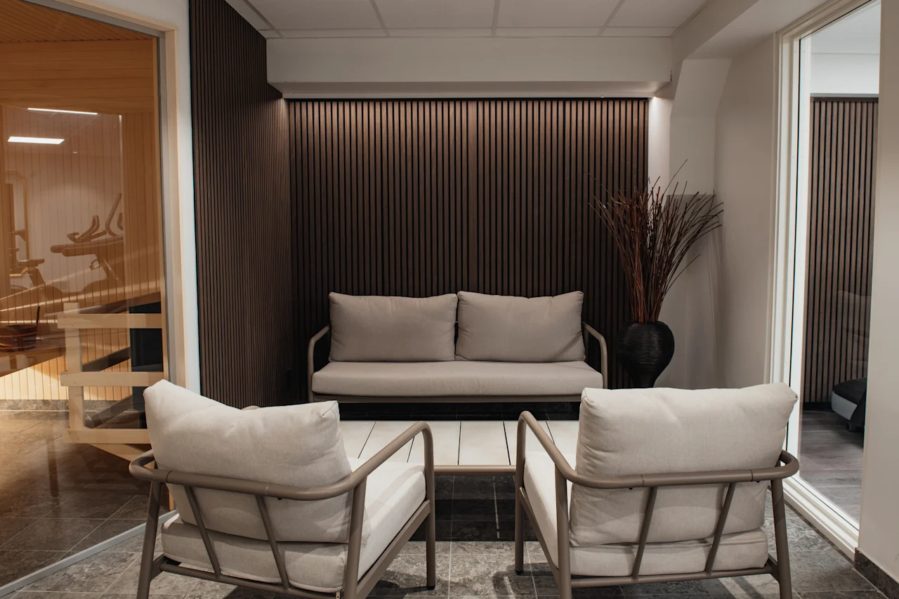 En modern loungeavdelning med bekväma sittplatser, ett soffbord och en dekorativ växt, intill ett glasinkapslat friskvårdsutrymme.