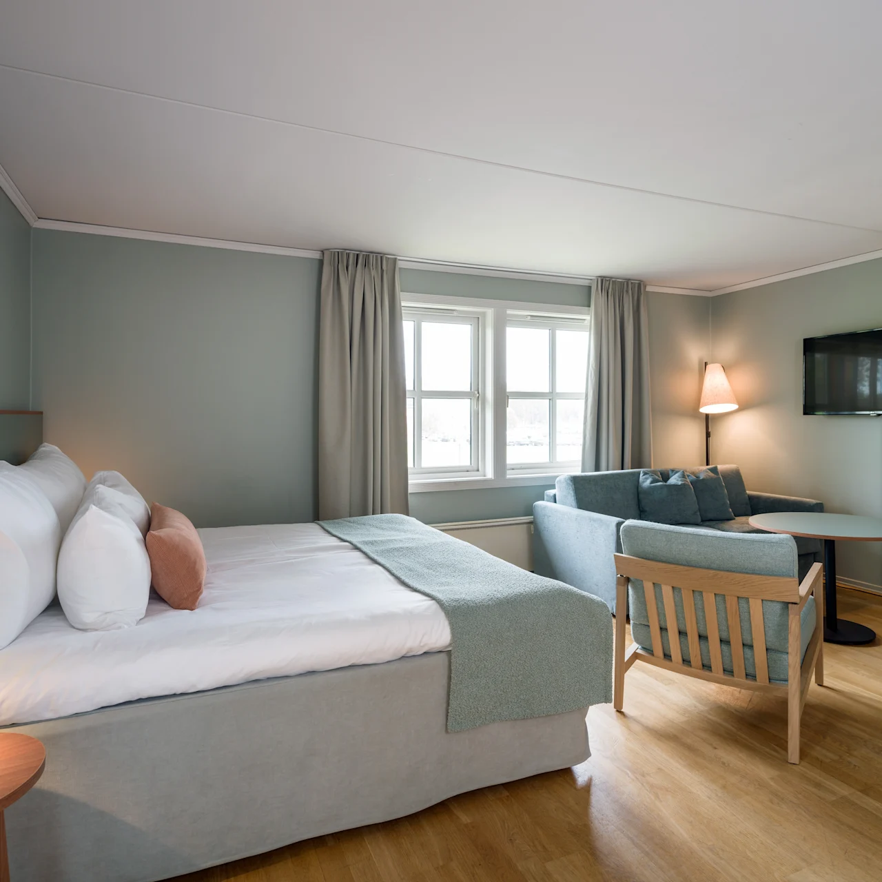 A neatly made bed with pillows and blanket, situated beside a small table and chair. A sofa and lamp are in the background near a window with curtains, in a cozy, softly lit room.