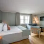 En pent redt seng med puter og teppe, ved siden av et lite bord og stol. En sofa og lampe er i bakgrunnen nær et vindu med gardiner i et koselig, mykt opplyst rom.