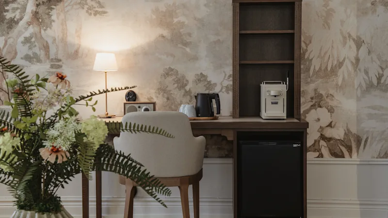 Et varmt og indbydende skrivebordsområde i et værelse med mønstret tapet, med lampe, elkedel og minikøleskab.