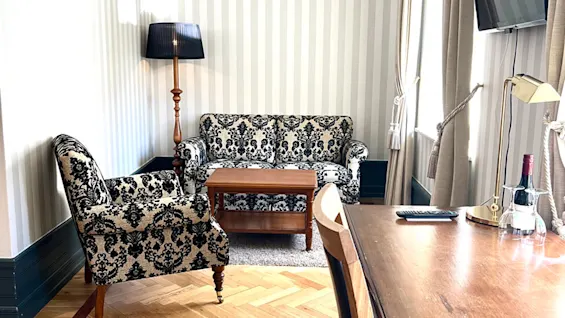 En mønstret lenestol og sofa står ved siden av et trebord under en stripet vegg og lampe, i et rom med tregulv og pult med en flaske og glass.