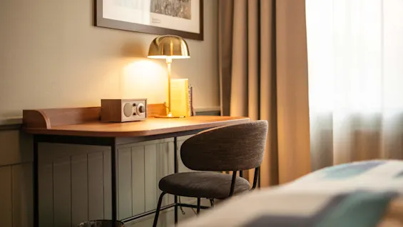 Ett träskrivbord med en vintage radio, böcker och en lysande lampa mot ett inramat foto. En grå stol står framför, med beige gardiner bredvid.