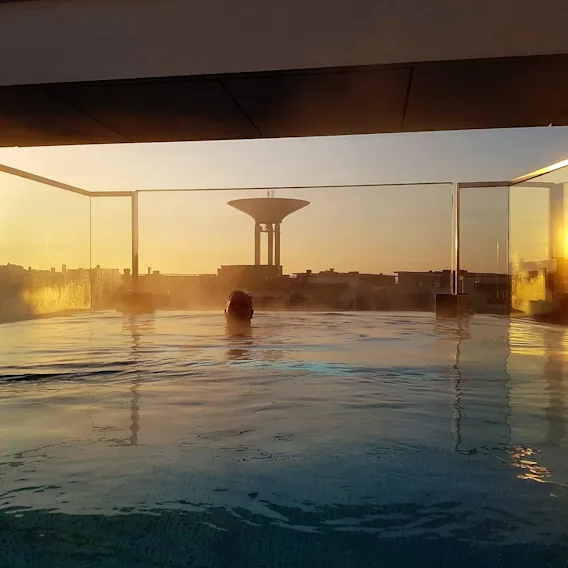 Quality Hotel™ Öresund – Rooftop Spa. En person kopplar av i en ångande takpool med en gyllene solnedgång över staden.