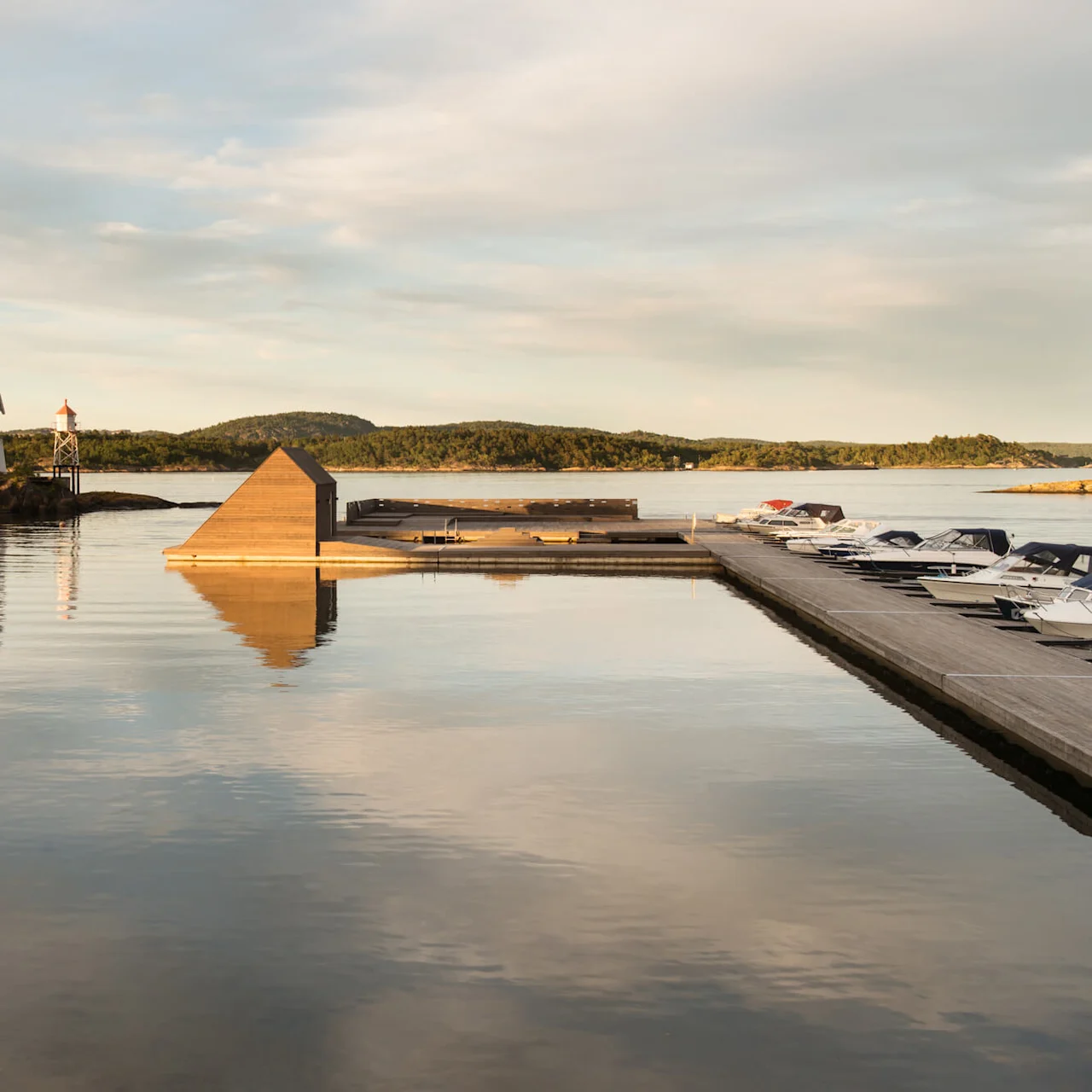 A wooden pyramid structure rests on calm water beside a dock with boats, surrounded by trees and distant hills under a cloudy sky.