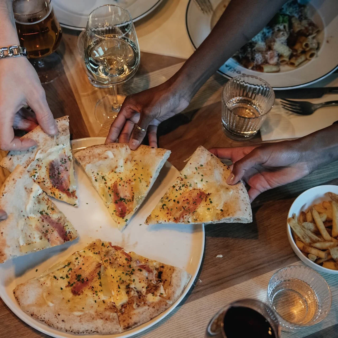 Pizza slice held by four hands, lifted from a plate with fries, drinks, and additional food dishes surrounding it on a wooden table. Various plates and a bottle are visible.