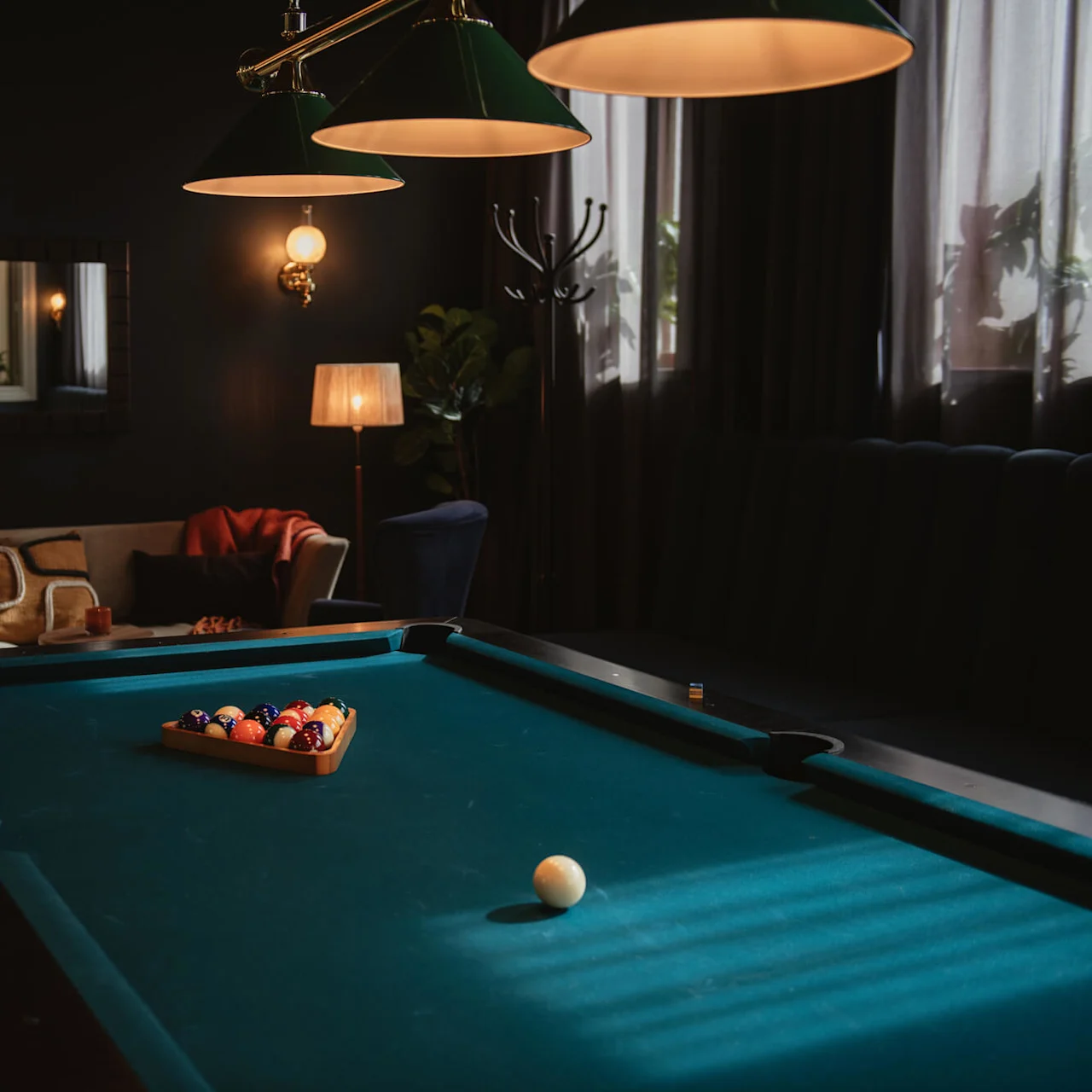 Pool table holds racked balls, unplayed, in dimly lit room with cozy seating, lamps, and wall mirror, evoking a relaxed, inviting atmosphere.