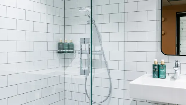 Ett glasvägg bashörna med en regnskydd i ett badrum med vita kakelplattor och en elegant handfat. Flera flaskor med toalettartiklar är prydligt ordnade på en hylla.