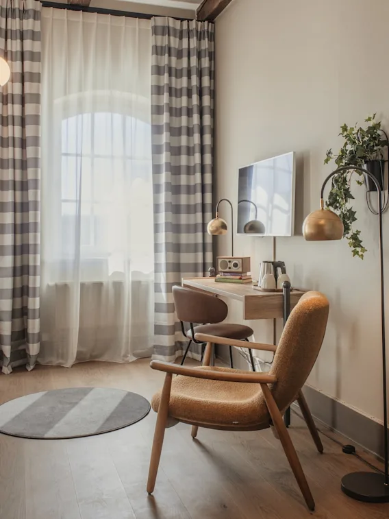 Viihtyisä hotellihuone, jossa sänky, työpöytäalue ja mukava nojatuoli, ruudulliset verhot ja lämmin valaistus.