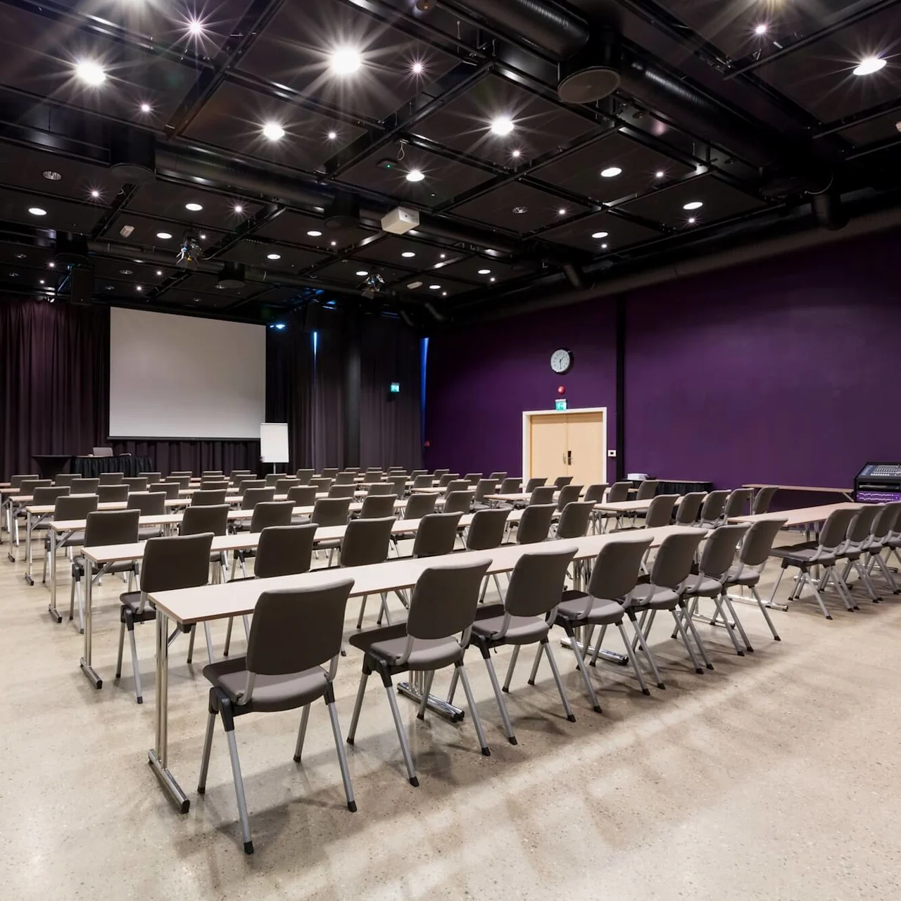 Rader av stolar och bord är vända mot en stor projektionsskärm i ett rymligt konferensrum med lila väggar och ett polerat betonggolv.