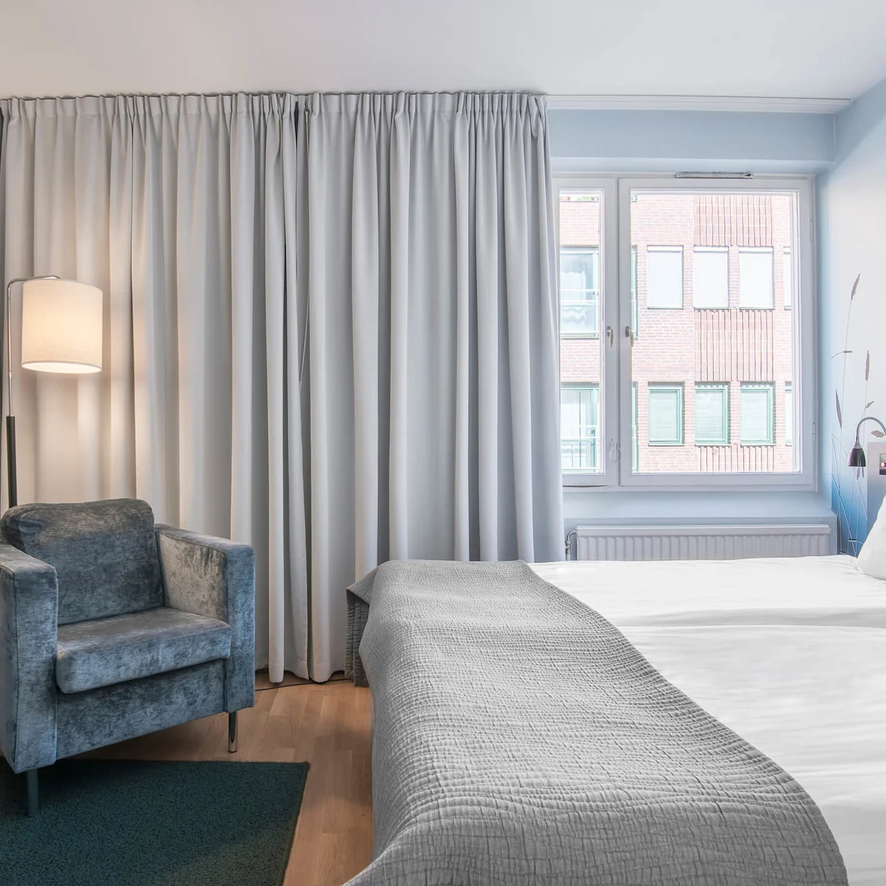 Ett mysigt hotellrum med en säng med vita sängkläder, en grå fåtölj, och en hängande lampa, beläget vid ett fönster med stängda gardiner. En väggmonterad TV finns.
