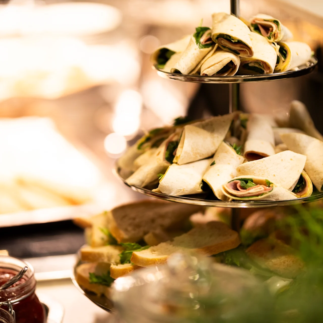 Layered platter hosting neatly rolled sandwiches, surrounded by other food items, located on a buffet table with jars of condiments nearby. Various trays and greenery enhance the presentation.