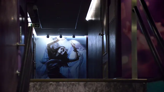A large poster of a person in a dynamic pose adorns the wall at the top of dimly lit stairs, surrounded by dark walls and ceiling lights.