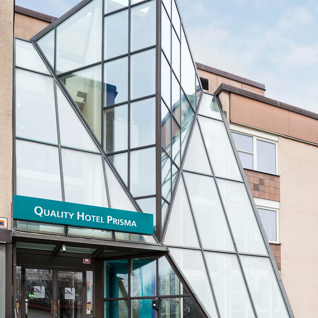 Glasingång med trekantiga paneler som reser sig från trappor, med skylten "QUALITY HOTEL PRISMA" i en modern byggnadskontext.