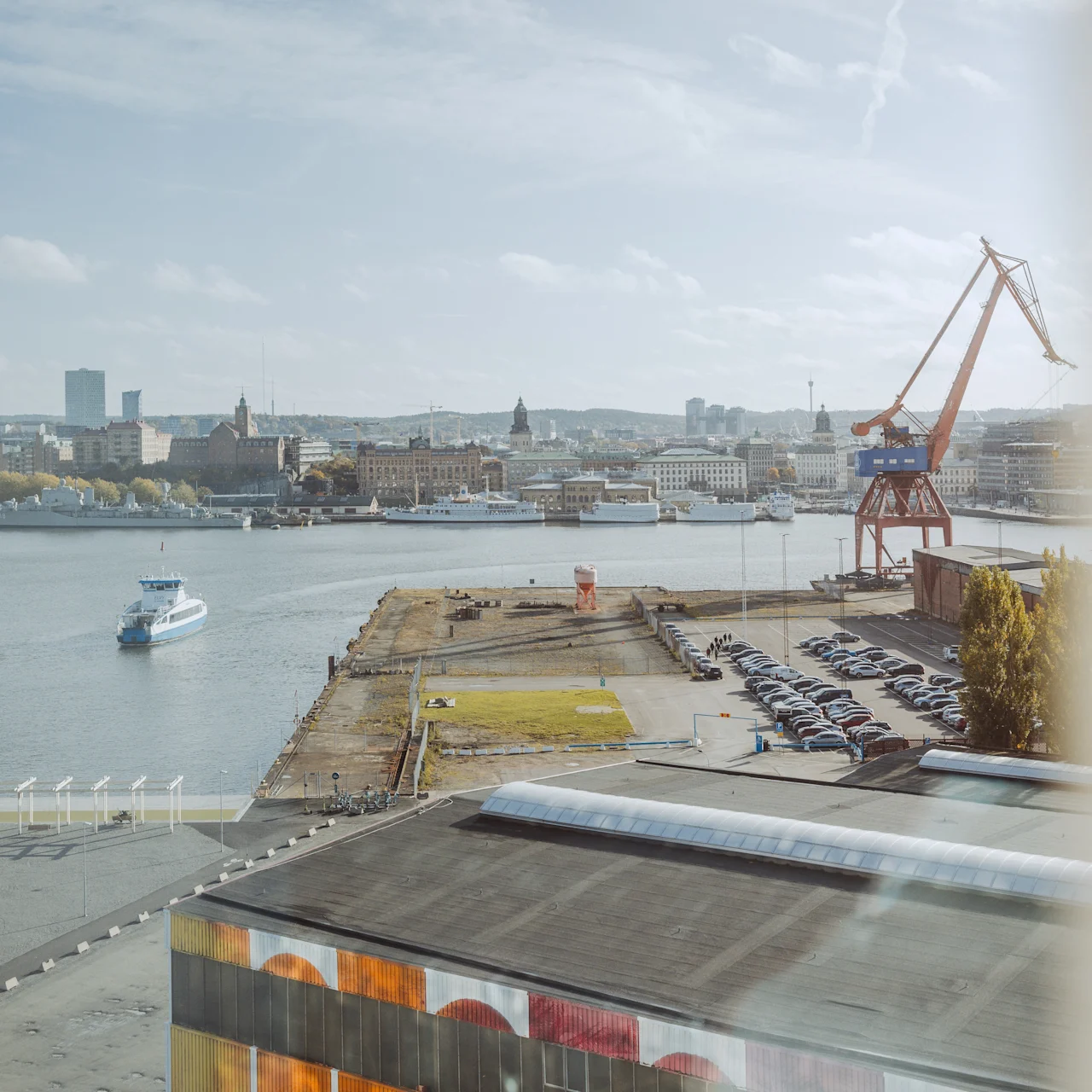 En flod med en båt som seglar; stor kran står vid kajen; industribyggnader och parkerade bilar synliga runtom; stadsbild med olika byggnader i bakgrunden under en delvis molnig himmel.