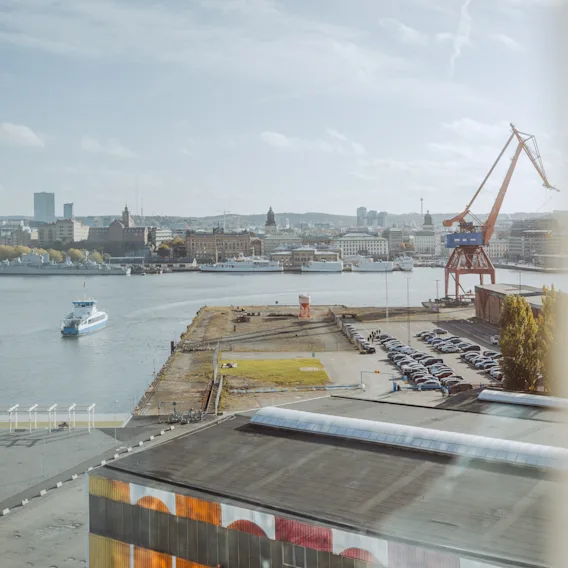 En elv med en båt som seiler; stor kran står på kaia; industribygninger og parkerte biler synlige rundt; bybilde med forskjellige bygninger i bakgrunnen under en delvis skyet himmel.