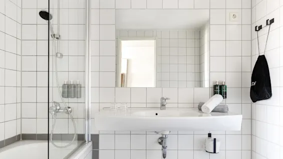 A white-tiled bathroom features a sink with folded towels and toiletries, a bathtub with a glass panel and a wall-mounted showerhead, creating a clean, minimalist look.