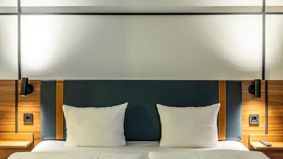 A close-up of a double bed with white bedding and two pillows. The headboard is blue with wooden panel details and orange accents. Reading lamps and small bedside tables are positioned on each side of the bed.