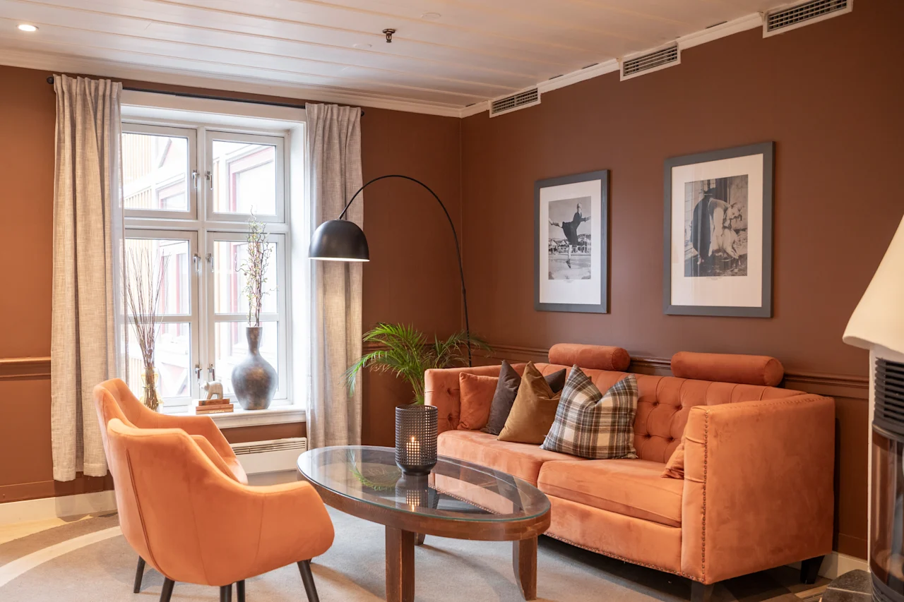 Oransje sofa og stol står i en koselig brunmalt stue, utsmykket med to innrammede bilder, en stålampe og et trebord, nær et vindu med lette gardiner.