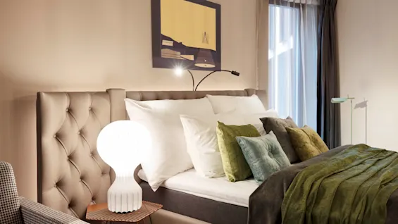 Bed adorned with white and green pillows sits beside a small side table holding a white lamp. A modern painting hangs above, while a window with sheer curtains illuminates the room.