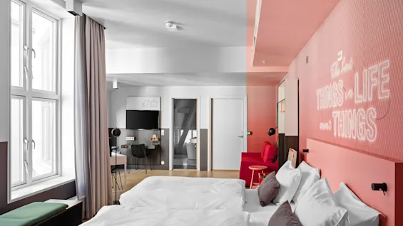 Hvit seng med puter står i et moderne rom med rosa vegger. Tekst på veggen lyder, "The best THINGS in LIFE aren't THINGS." Et skrivebord og TV er nær et vindu.