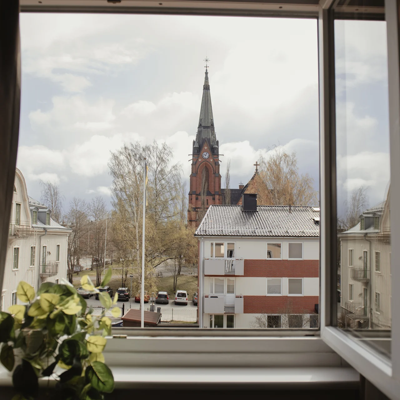 A church tower stands tall outside an open window, surrounded by trees and residential buildings. A potted plant rests on the windowsill.