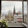 A church tower stands tall outside an open window, surrounded by trees and residential buildings. A potted plant rests on the windowsill.