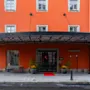En levende orange bygning med store vinduer har en overdækket indgang med en rød løber. Potteplanter flankerer døren, og skilte siger "hotel." Det ligger på en brostensbelagt gade.