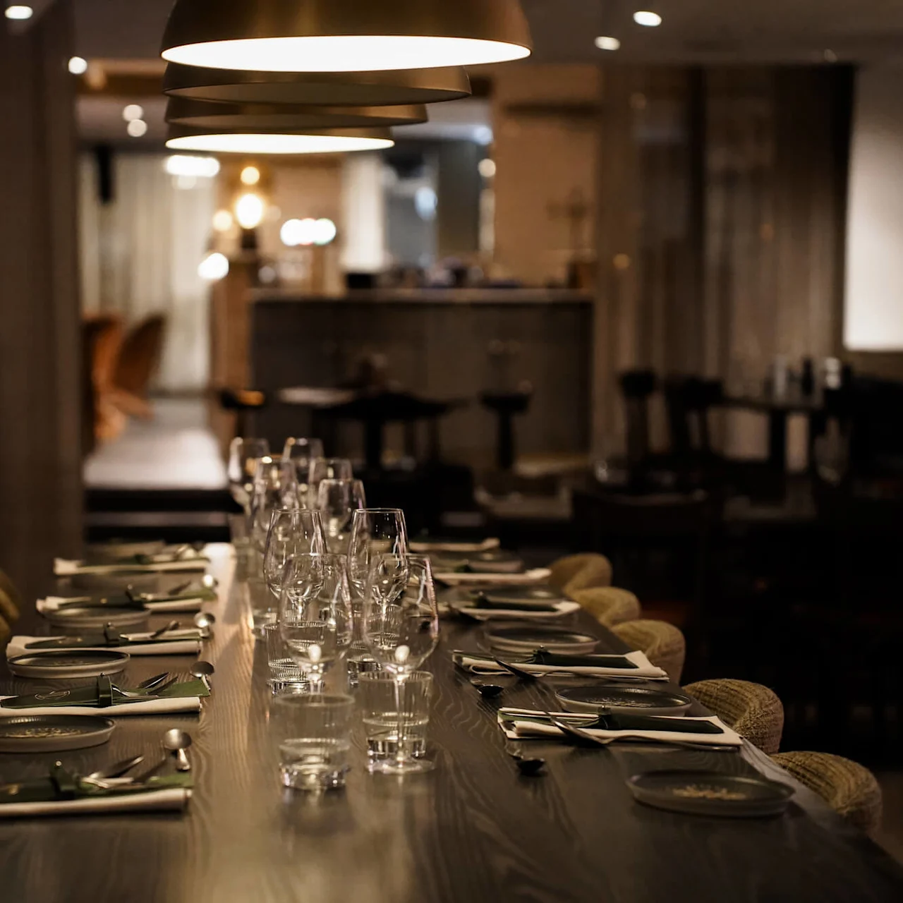Ett långt matbord är dukat med tallrikar, glas och bestick, belyst av taklampor, i en svagt upplyst restaurang med en mysig atmosfär.