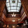Grand chandelier hanging from an ornate stained glass ceiling in a luxurious, wood-paneled room with elegant furnishings, framed portraits, and a large fireplace. 