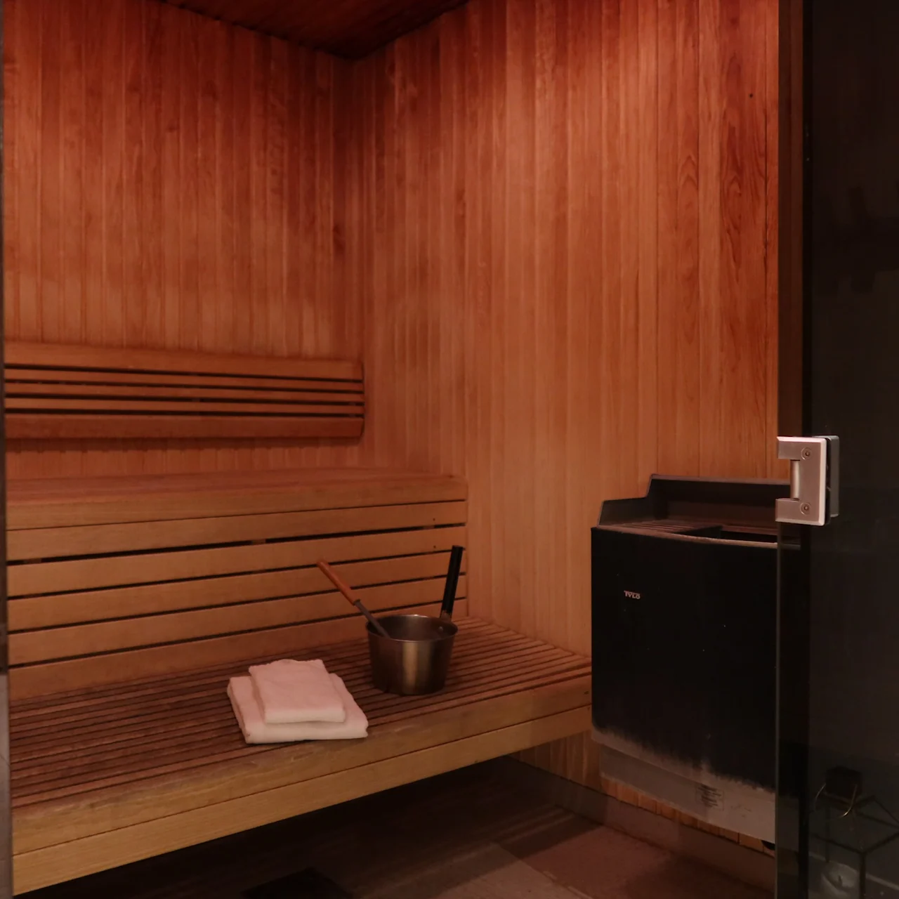 Wooden bench holds a metal bucket and folded towels; set in a warm-toned sauna room with wood-paneled walls and a glass door.