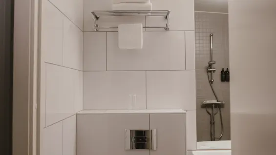 Valkoinen wc-istuin keskellä metallihyllyn, jossa pyyhkeitä, alla. Vieressä moderni suihkupää ja pesuallas laatoitetussa kylpyhuoneessa.