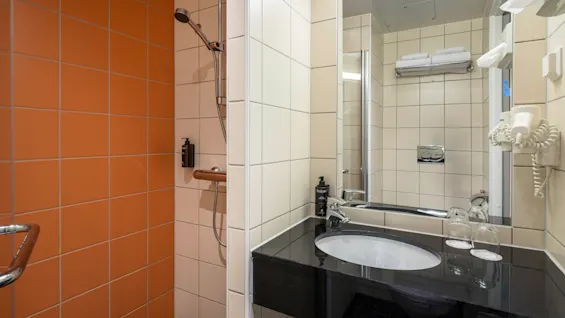 Et moderne badeværelse har en sort bordplade med vask, ved siden af en bruseniche med orange fliser. Rummet indeholder en vægmonteret hårtørrer og pænt stablede håndklæder.